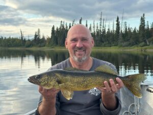 Person holds a large fish in a boat on a lake surrounded by trees under a cloudy sky.