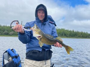 Person in a blue jacket holds fish with grip on a boat with forest and cloudy sky background.