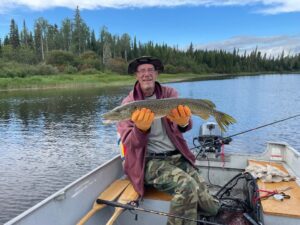Man in a boat holds large fish on a lake, with forested shoreline and partly cloudy sky.