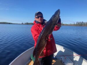 Person in red jacket holding large fish on a boat, lake and trees in background.