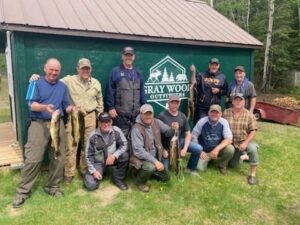 Eleven people pose in front of green building, some holding fish, grassy and wooded background.