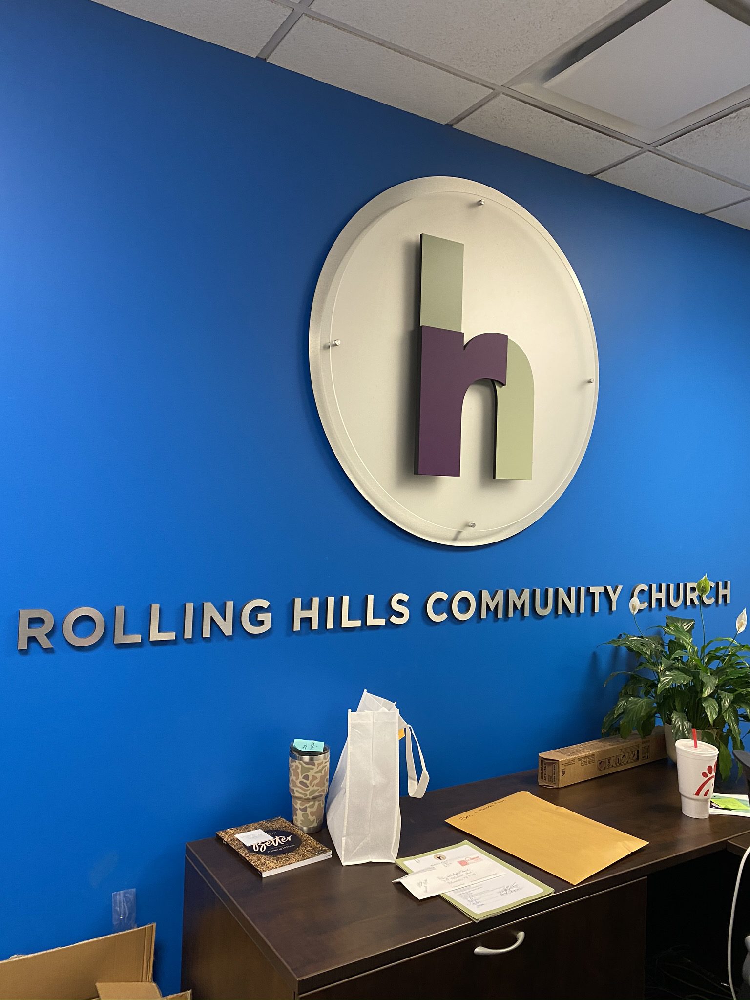 Blue wall with "Rolling Hills Community Church" sign, logo; desk holds items like a potted plant.