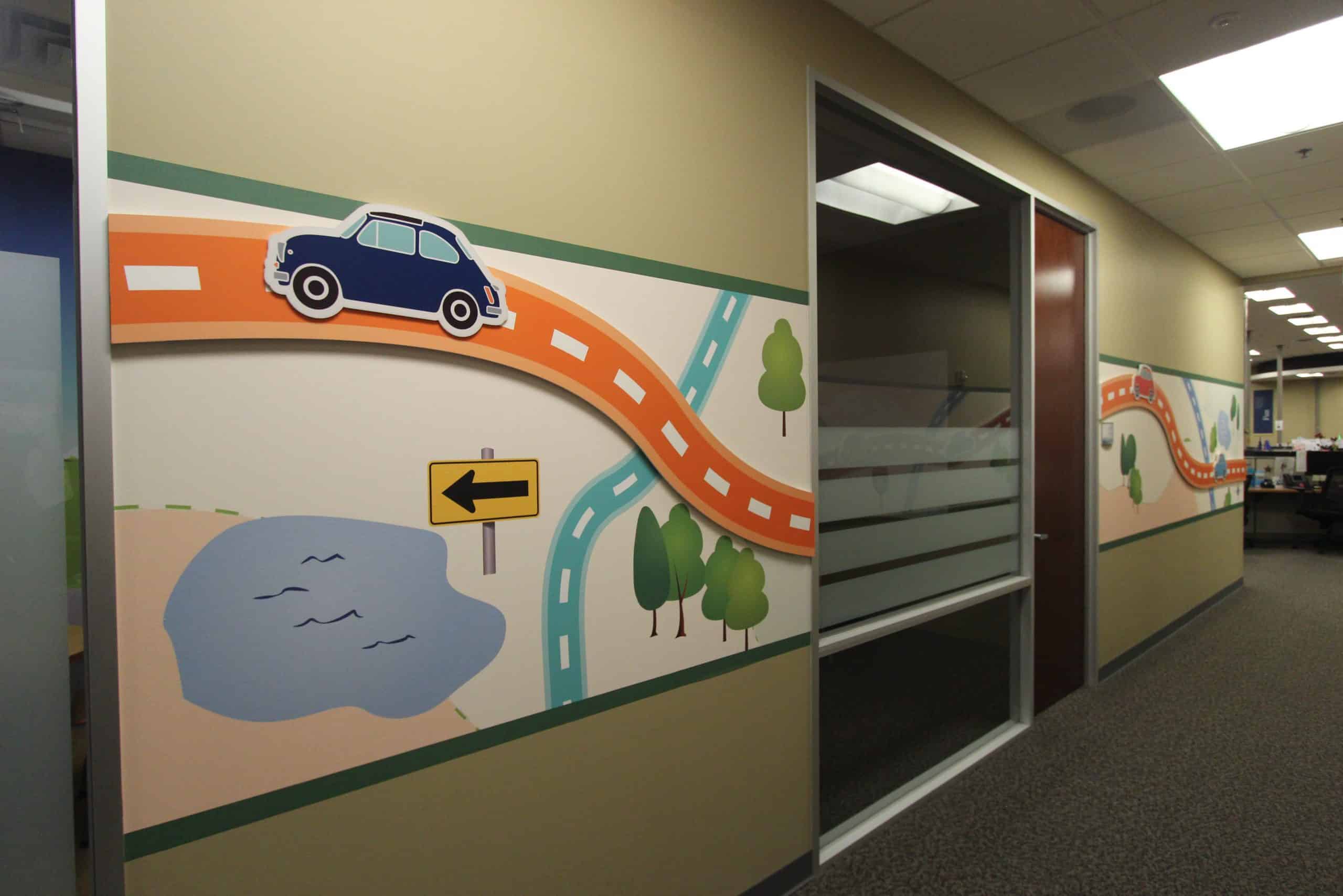 A fun custom interactive display in front of the human resources department features a background made of adhesive wallpaper, with CNC-cut, directly printed PVC cars and roads mounted away from the wall, and a hatch that allows the cars to move along the road.