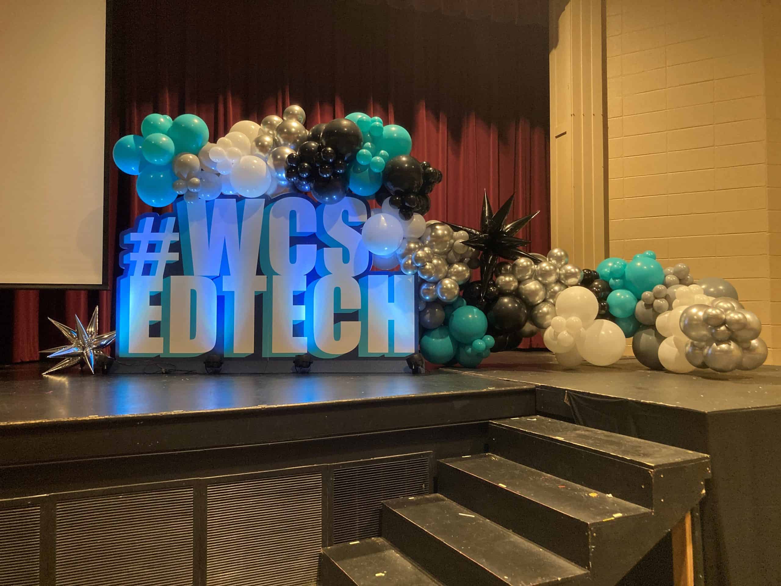 Stage with 3D cut gator board Standee "#WCS EDTECH" sign, colorful balloons, maroon curtains, and silver star decoration.